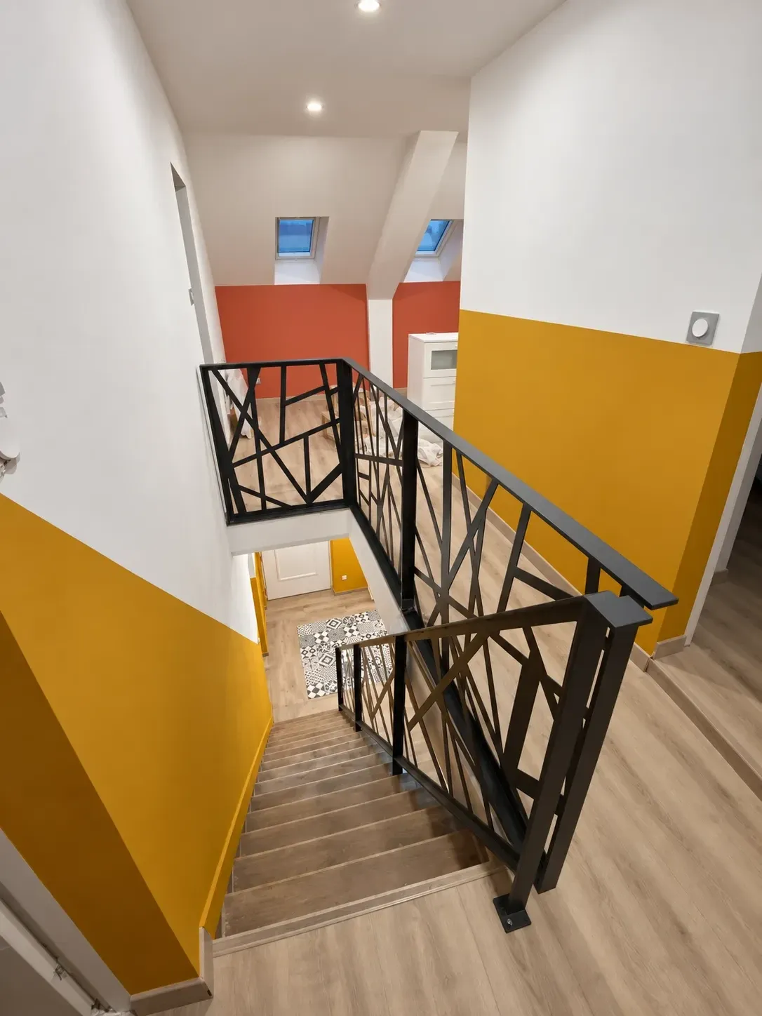 Palier d'étage sous combles rénové avec garde-corps métallique à motifs géométriques, murs jaune moutarde et terracotta
