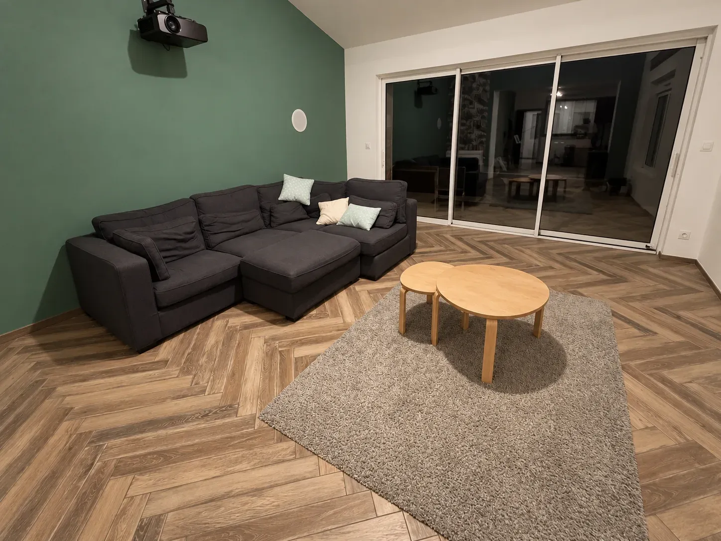 Salon cinéma aménagé avec canapé d'angle, vidéoprojecteur, mur vert et carrelage effet parquet à chevrons